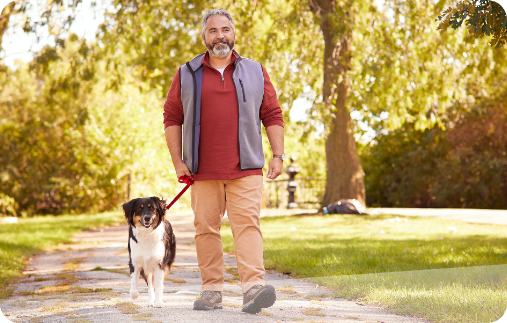Photograph of a man walking his dog on a park path on a beautiful day.